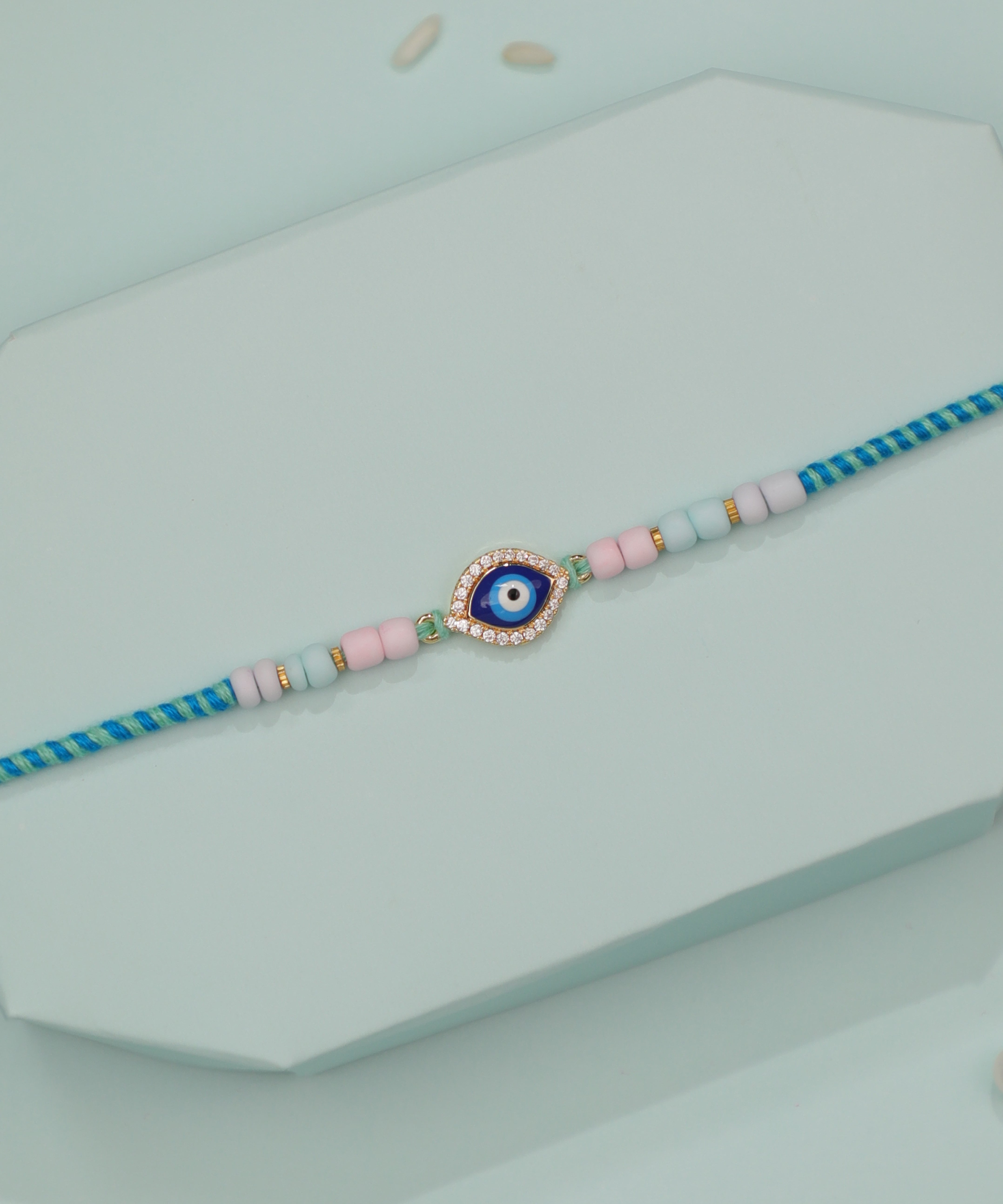 Gold-tone Rakhi with evil eye charm, blue enamel, CZ stones, and pastel pink and blue beads on braided thread.