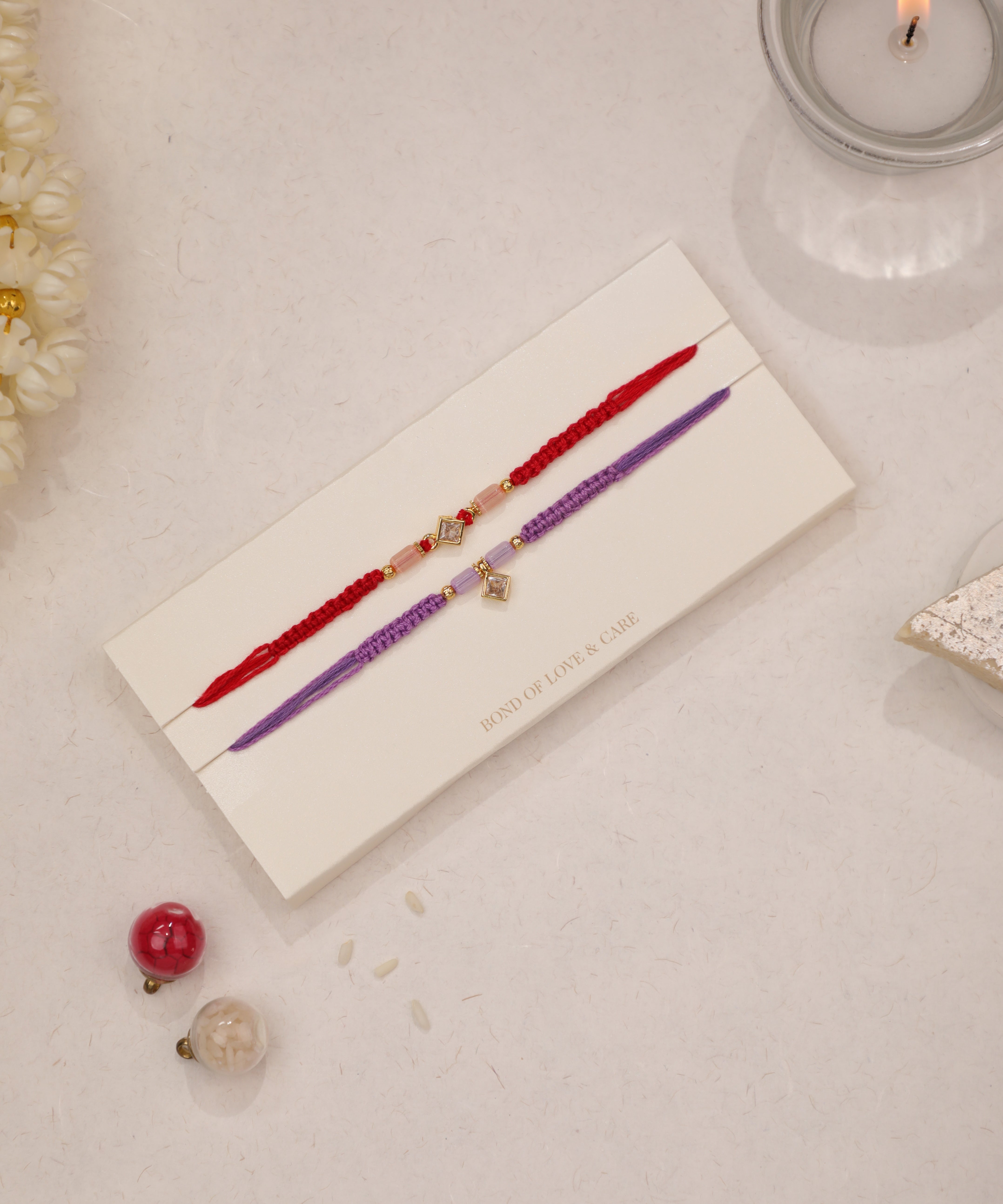 Gold-tone Rakhi with clear stones and pink beads on red thread, and purple thread with square charm