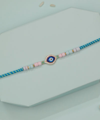 Gold-tone Rakhi with evil eye charm, blue enamel, CZ stones, and pastel pink and blue beads on braided thread.