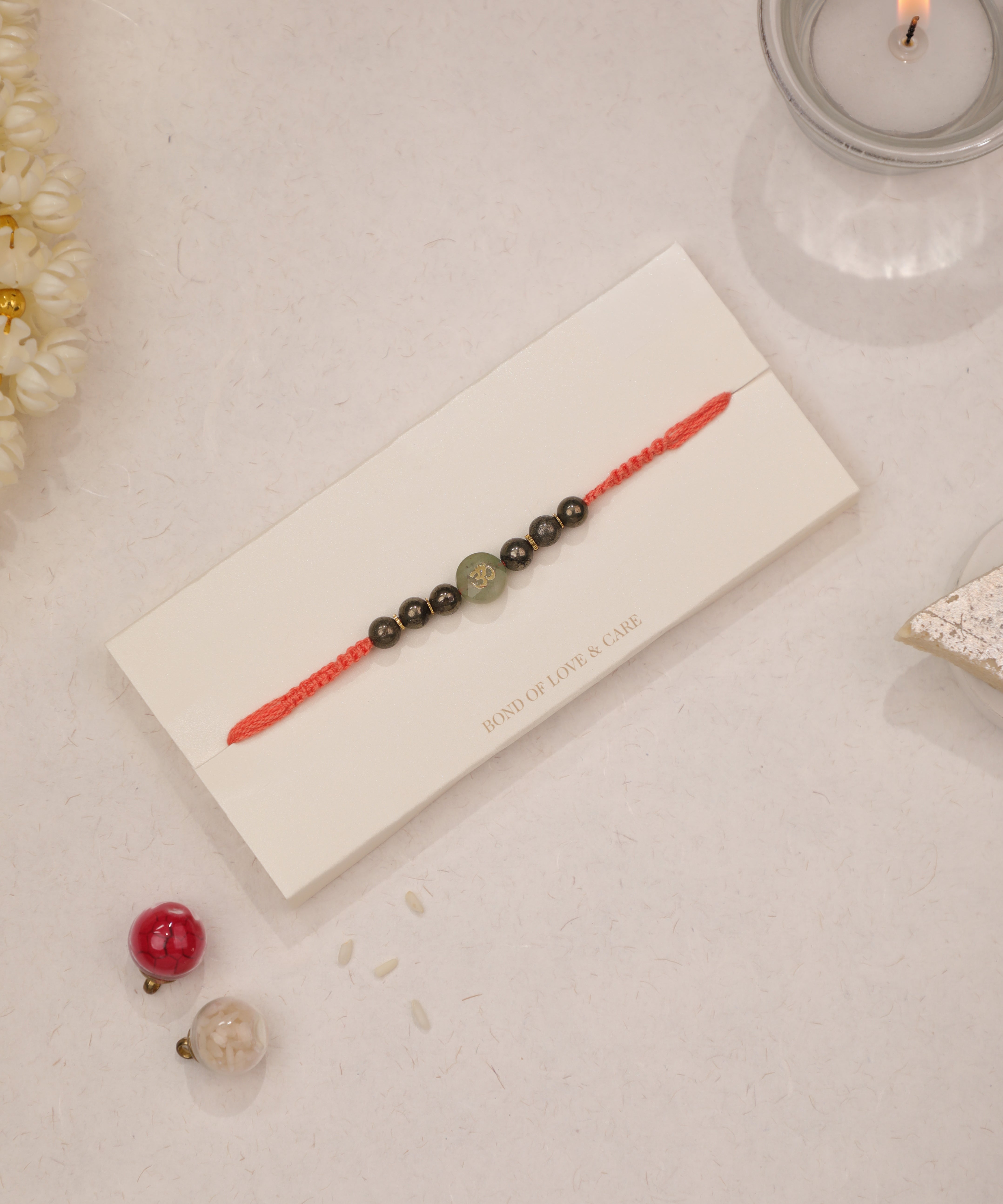 Red braided Rakhi with central green Om symbol bead and dark metallic beads.