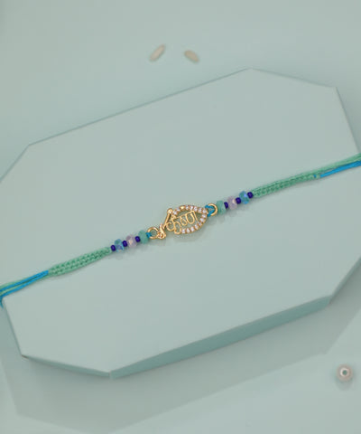 Gold-tone Rakhi with leaf-shaped Krishna script charm and blue-green braided string with colorful beads.