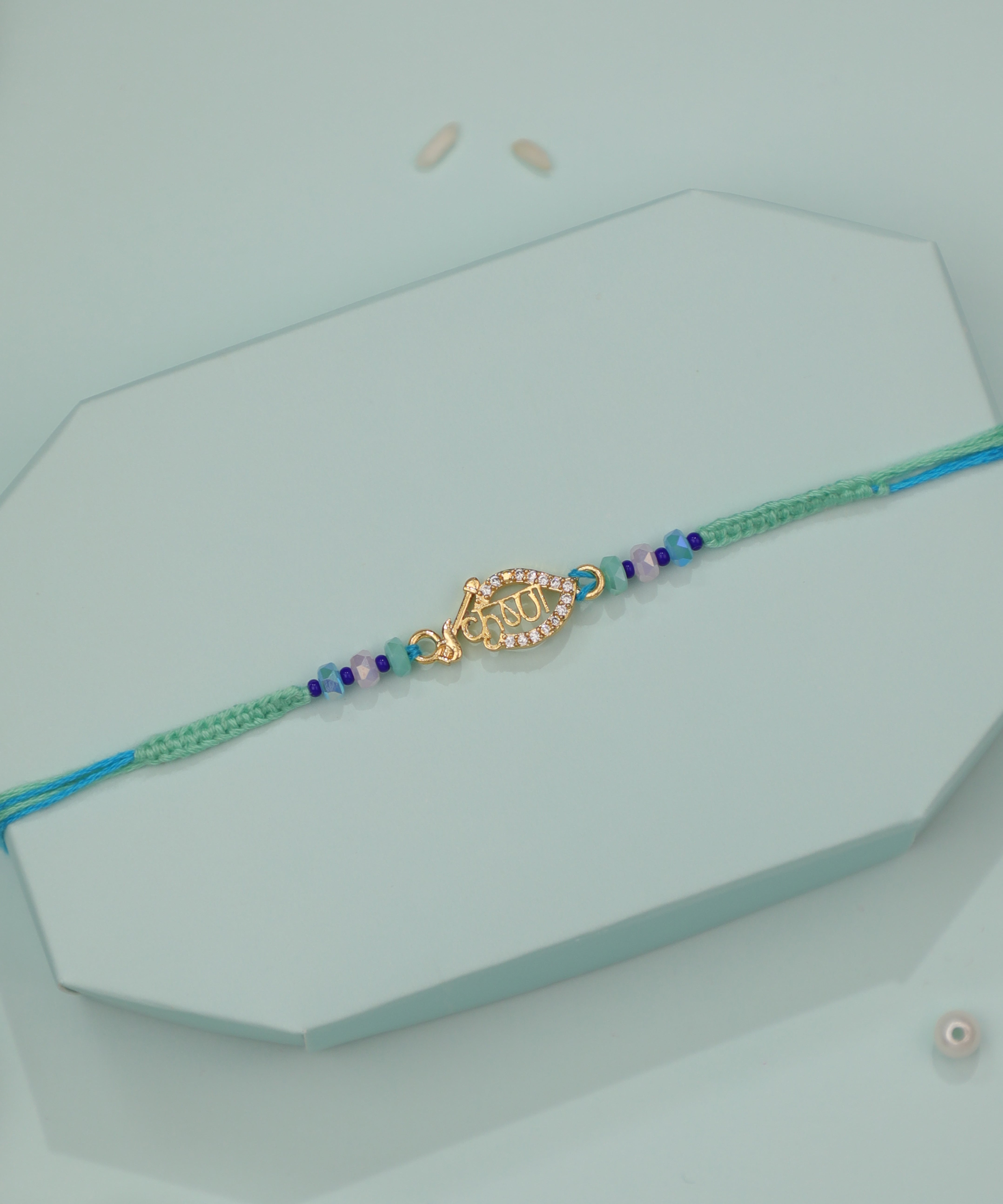 Gold-tone Rakhi with leaf-shaped Krishna script charm and blue-green braided string with colorful beads.