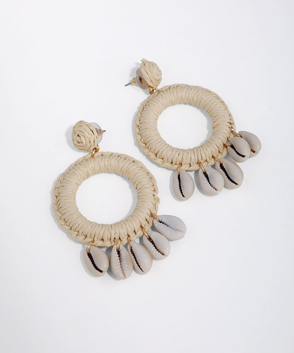 Gold-tone earrings with large raffia-wrapped hoops and dangling cowrie shells on a white background.