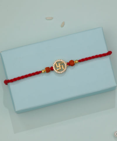Gold-tone Rakhi with Swastika design, clear stones, and rudraksha beads on a red thread