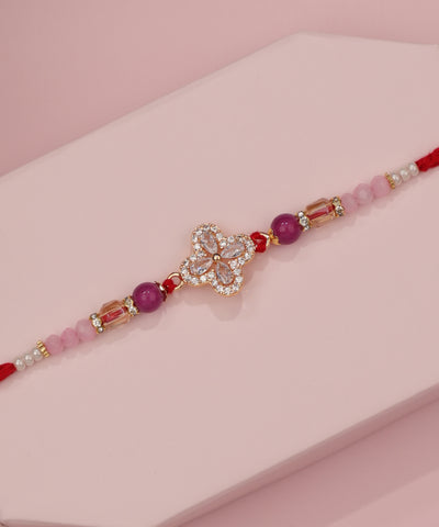 Gold-tone Rakhi with sparkling clover motif, purple and pink beads, and red thread ties.